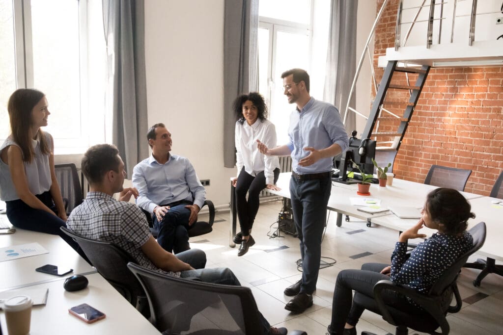 Eine Gruppe von sechs Personen in einem Besprechungsraum diskutiert über Arbeits- und Organisationspsychologie, wobei eine Person als Sprecher steht. Der Raum hat Ziegelwände und einen Tisch.