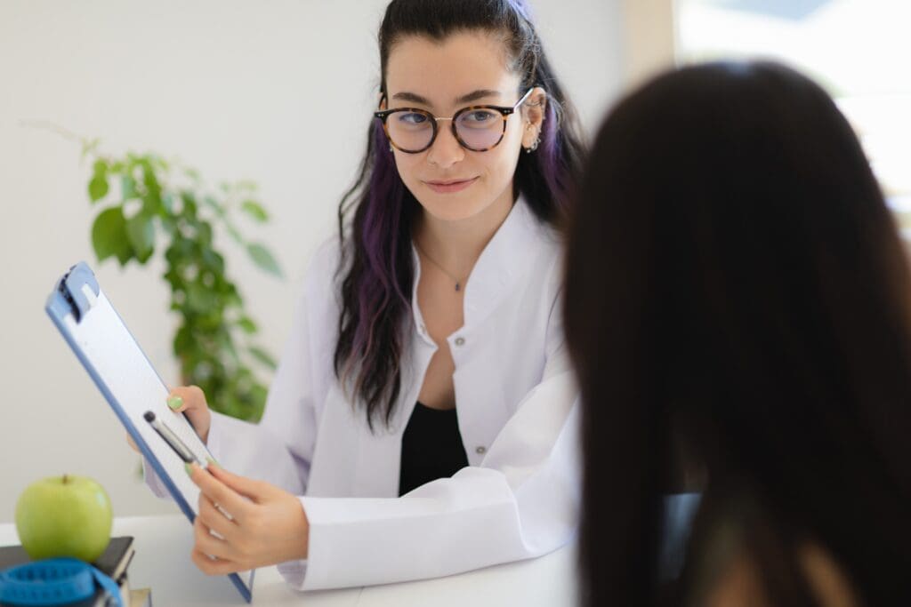 Eine Studierende der Gesundheitspsychologie mit Brille und einem weißen Kittel zeigt einem anderen Menschen etwas auf einem Klemmbrett.