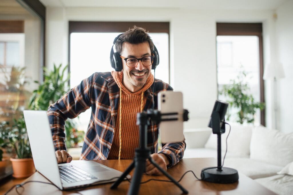 Ein Studierender wendet seine Kenntnisse in Kommunikations- und Medienpsychologie an, indem er einen Podcast macht. Notebook, Smartphone und Mikrofon sind auf seinem Schreibtisch verteilt und er lächelt entspannt.