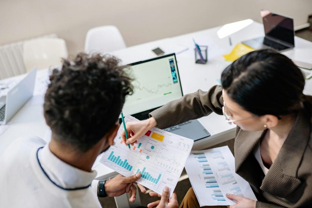 Eine junge Frau in Business-Kleidung zeigt einem jungen Mann ein Diagramm in ihren Unterlagen. Auf dem Tisch neben den beiden steht ein Notebook mit weiteren Diagrammen, im Hintergrund sind weitere Arbeitsmaterialien zu sehen.