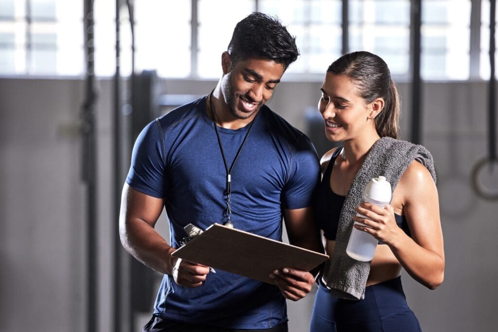 Ein Mann und eine Frau in einem Fitnessstudio, die über Sportpsychologie und Trainingspläne sprechen, lächelnd und eine Klemmbrett sowie eine Wasserflasche haltend.