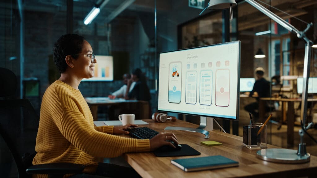 Eine Person arbeitet an einem Projekt in Wirtschaftspsychologie. Sie sitzt dafür vor einem großen Monitor, neben Notizbuch, Schreibtischlampe, Kaffeetasse. Im Hintergrund sind weitere Personen zu sehen, die in ähnlichen Settings arbeiten. Die Atmosphäre ist ruhig, konzentriert.