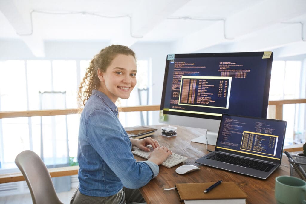 Eine junge Spezialistin für Künstliche Intelligenz (KI) arbeitet lächelnd an Notebook und Monitor.